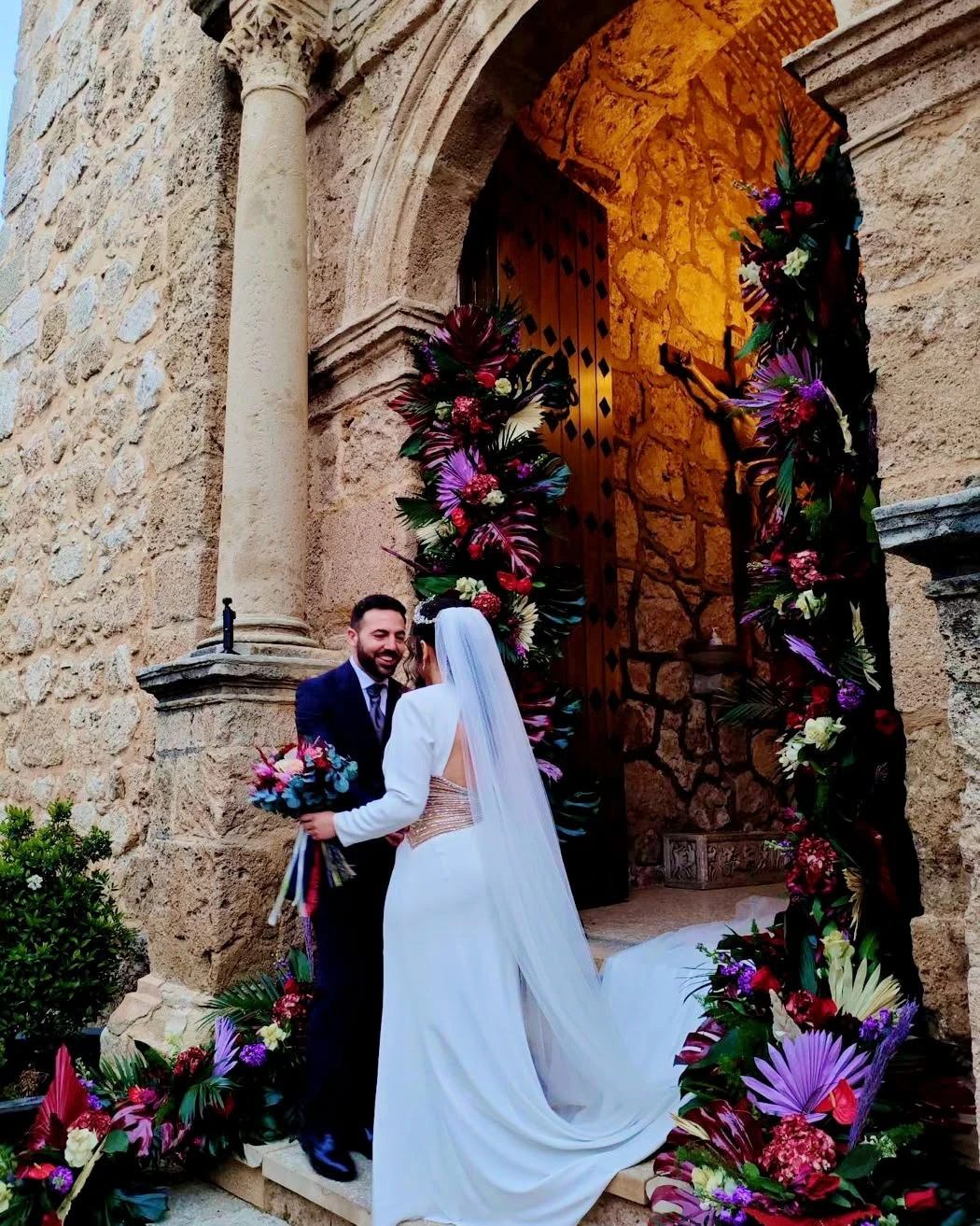 Novios en la puerta de la iglesia, decorada con flores en tonos morados, rojos y verdes